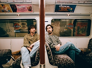 Two men sit apart on a subway train, facing each other with neutral expressions. Advertisements are visible above them and a pole divides the scene.