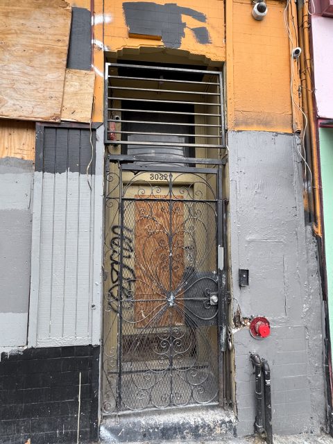 Wrought iron door with graffiti and a lock in front of a weathered, patched building facade, address number 303 on doorframe, wires and fire hydrant visible nearby.
