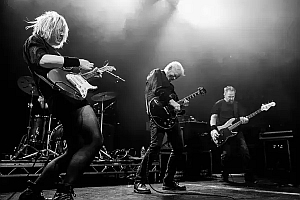 Three musicians play electric guitars and bass on stage under dramatic lighting, with drums visible in the background.