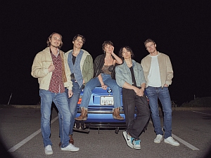 Five people stand and sit on the back of a blue car in a parking lot at night, posing for the photo.