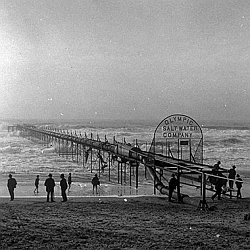 Black and white photo of people standing near a long wooden pier extending into the ocean, with a sign reading "Olympic Salt Water Company.