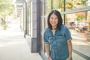 A woman wearing a denim shirt stands smiling on a sunny sidewalk beside a building with large windows.