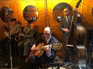 Four musicians in suits perform indoors; one plays guitar, one upright bass, one saxophone, and one trumpet. Circular mirrors are on the wall behind them.