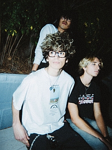 Three young people sit and pose outdoors at night; one wears glasses and a white shirt, another has a black shirt, and the third stands behind them.