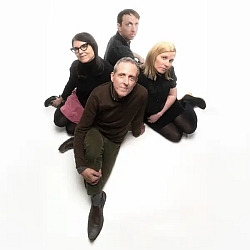 Four people sit on the floor in a white studio, arranged in a loose diamond shape, facing the camera with neutral expressions.