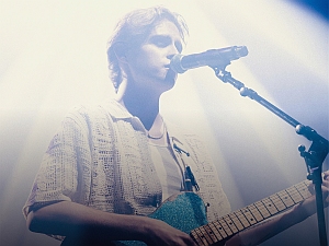 Person playing electric guitar and singing into a microphone on stage, illuminated by bright, white stage lights.
