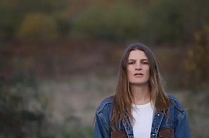 A person with long brown hair wearing a denim jacket and white shirt stands outdoors with a blurred natural background.