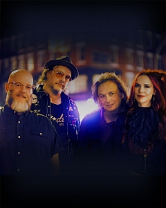 Four adults stand close together, posing for a group photo in front of a blurred, dimly lit background.