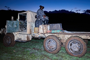 A person sits on the cab of a worn, mud-covered truck parked on grassy ground at dusk with mountains in the background.