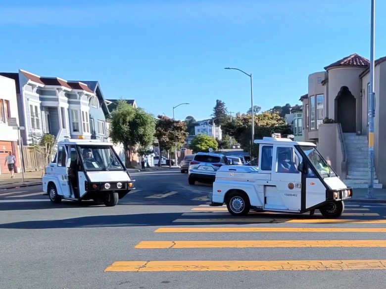 Two white parking enforcement vehicles drive through a residential intersection with houses, trees, and a yellow crosswalk visible.