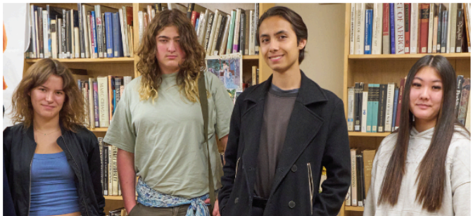 Four young people stand in front of bookshelves in a library, facing the camera and posing together.