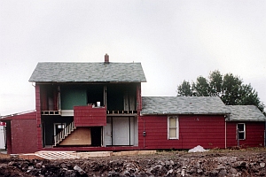 A red two-story house partially cut open, exposing interior rooms and staircase; construction debris and dirt are visible in the foreground.