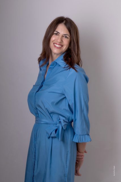 A woman with long brown hair, wearing a light blue belted dress, stands in front of a plain background and smiles at the camera.