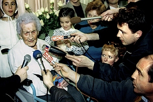 An elderly woman sits in a wheelchair surrounded by reporters holding microphones and recording devices, with several onlookers nearby.