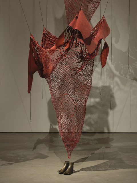 A suspended red mesh and fabric sculpture is anchored to the floor by two bronze legs and attached to the ceiling and walls by thin cords.