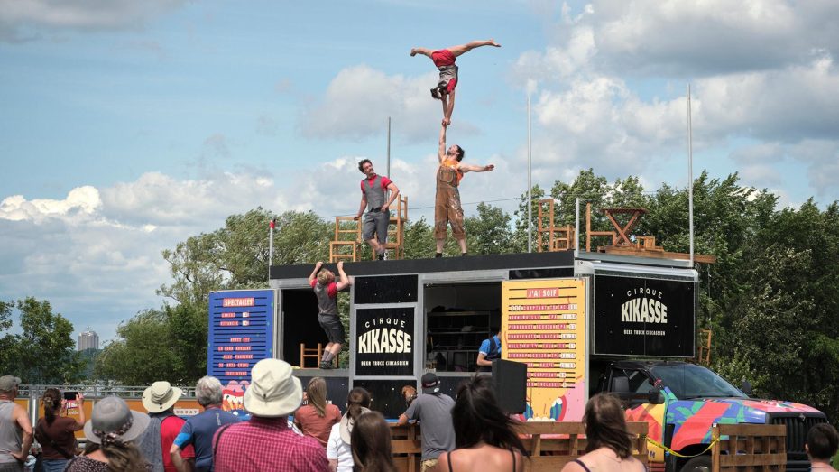 A circus performance on an outdoor stage features acrobats balancing and performing stunts, while a crowd watches under a partly cloudy sky.