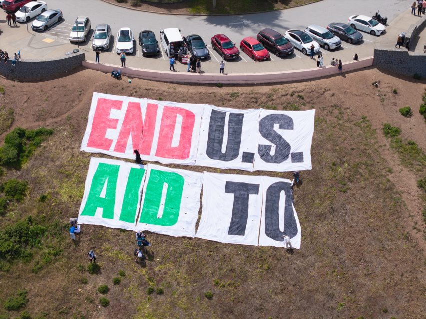 Large banners on a hillside read "END U.S. AID TO." Several people are around the banners, with parked cars and others standing along a nearby road above.