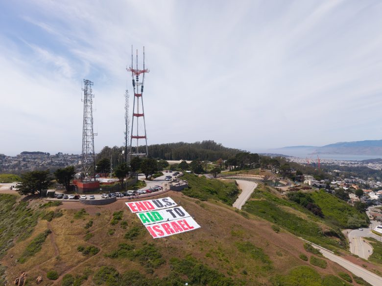 A hillside displays a large banner reading "END U.S.AID TO ISRAEL" near radio towers, overlooking a cityscape and coastline.