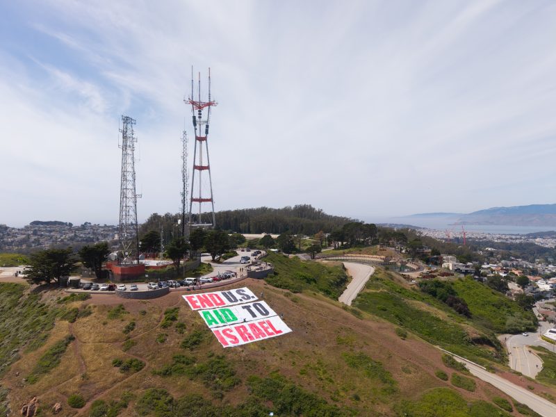 A hillside displays a large banner reading "END U.S.AID TO ISRAEL" near radio towers, overlooking a cityscape and coastline.
