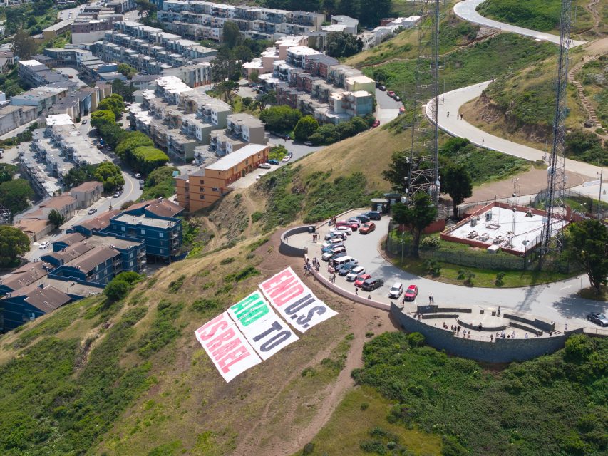 A large banner reading "END U.S. AID TO ISRAEL" is displayed on a hillside near a parking area, with people standing nearby and residential buildings in the background.