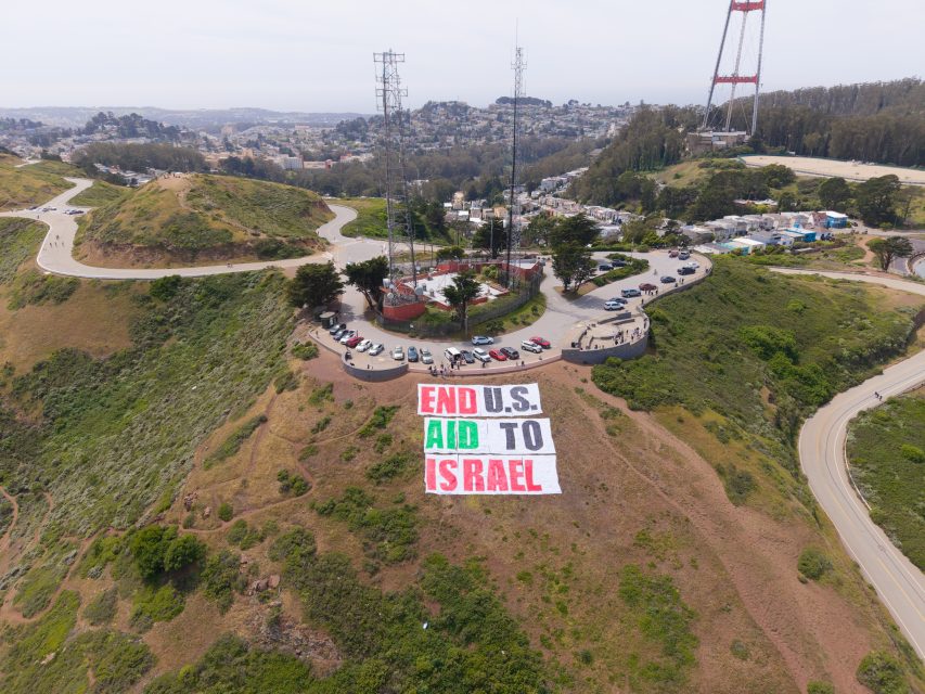 A large banner on a hillside reads "END U.S. AID TO ISRAEL" in red, black, and green letters, with roads, cars, and a cityscape visible in the background.