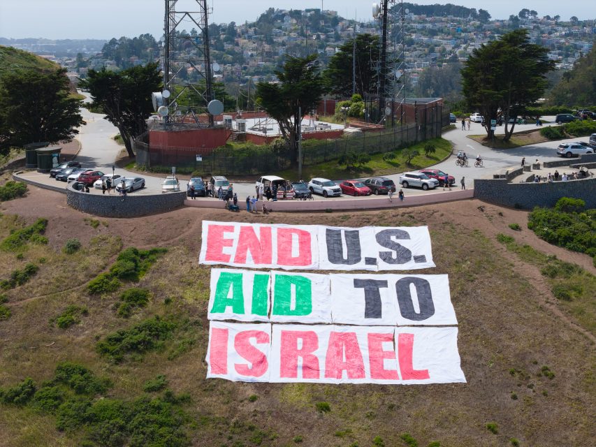 Large banners on a hillside spell out "END U.S. AID TO ISRAEL" near a road with parked cars, people, and telecom towers in the background.