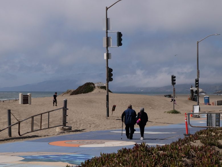 Two people walk along a painted path near a sandy beach, with traffic lights, some vegetation, and cloudy skies in the background.