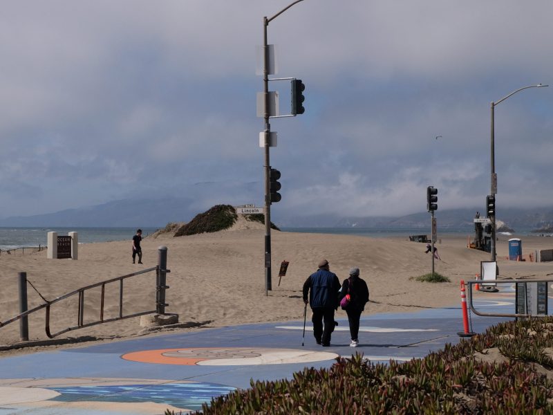 Two people walk along a painted path near a sandy beach, with traffic lights, some vegetation, and cloudy skies in the background.