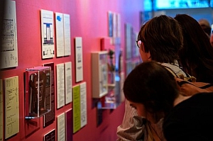 Several people closely examine documents and displays mounted on a pink wall in what appears to be an exhibition or gallery setting.