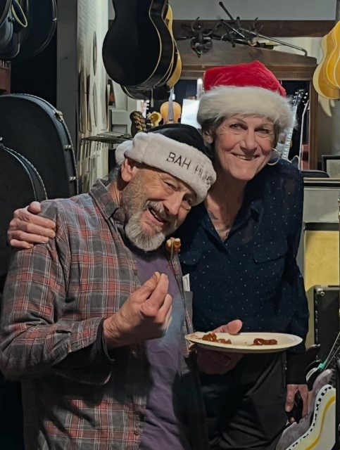 Two people wearing festive hats stand close together, one holding a plate of food and smiling, the other with an arm around him; guitars and cases are visible in the background.