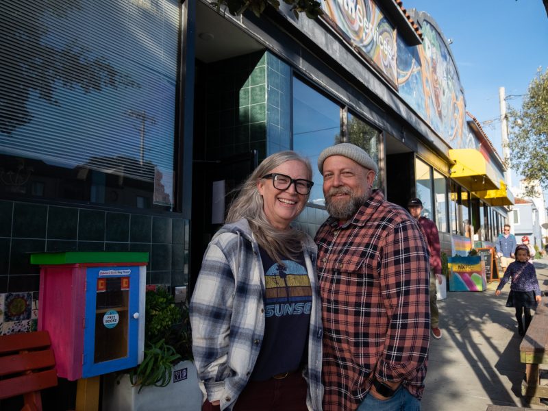 Two people stand close together and smile outside a colorful building on a sunny day. There are a few other people and painted decorations visible in the background.