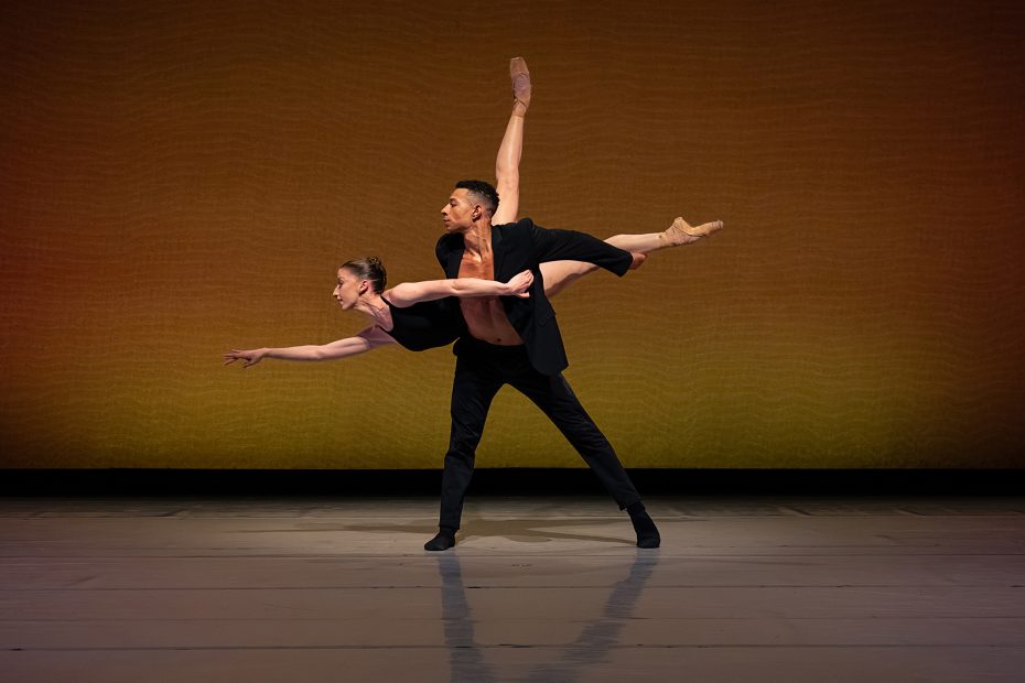 Two ballet dancers perform on stage; the male dancer supports the female dancer who is balanced on one leg with her other leg extended high behind her.