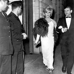 A woman in an evening gown and feathered shawl walks with a man in a tuxedo, passing two uniformed officers near a building entrance.