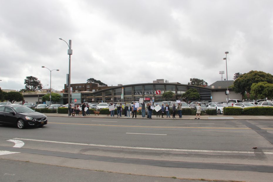 A group of people holding signs stands on the sidewalk outside a Safeway store on a cloudy day, with cars parked in the lot and driving by.