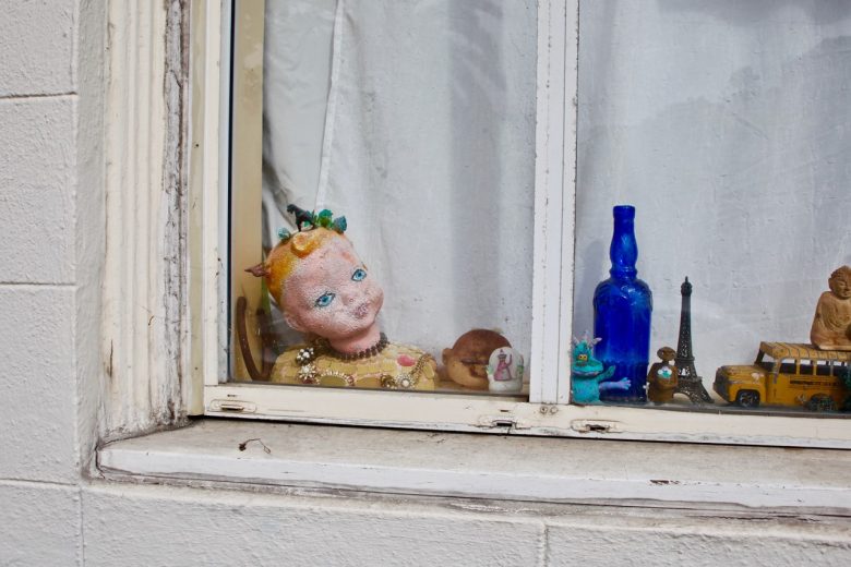 A display on a windowsill features a doll's head, a blue glass bottle, small figurines, a miniature yellow bus, and other assorted objects in front of a white curtain.