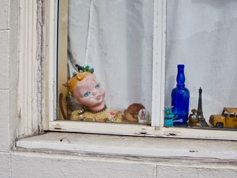 A window display with a damaged doll head, small figurines, a blue bottle, a yellow toy car, and a white curtain in the background.