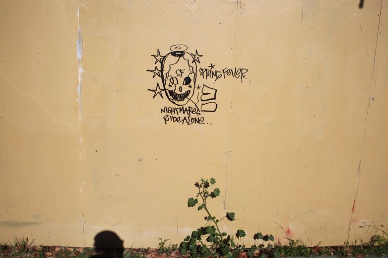 Graffiti of a skull with a halo and stars, text reads "Spring Fever" and "Nightmares Ride Along" on a yellow wall with a plant and shadow at the bottom.