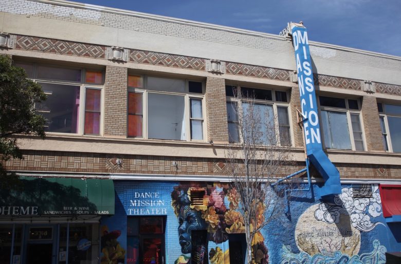 A building with a blue vertical "MISSION" sign, colorful mural, and a storefront reading "Dance Mission Theater" beneath second-story windows.