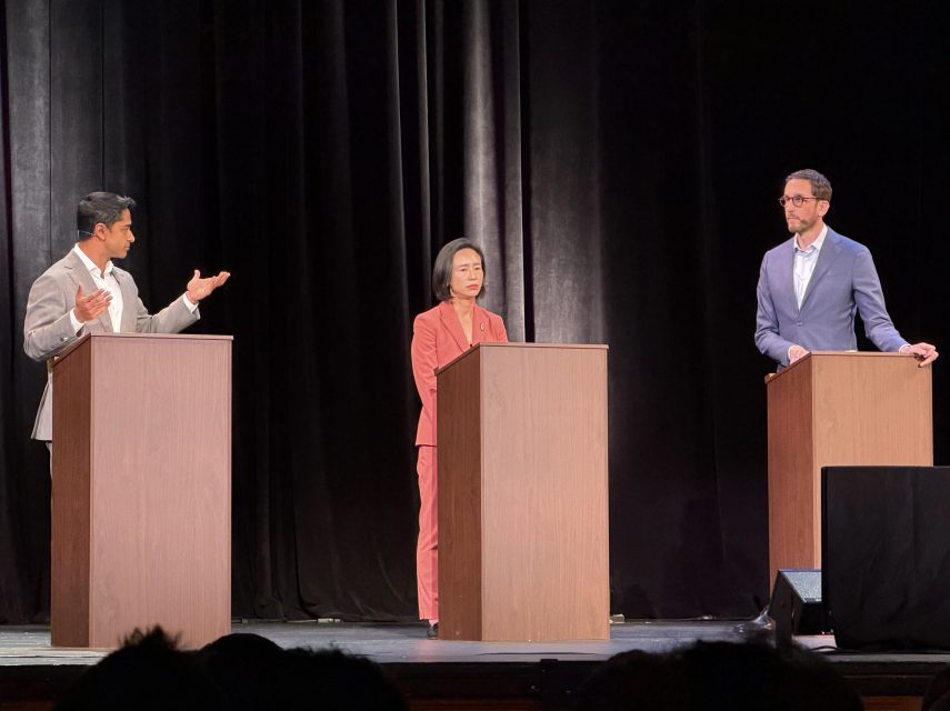 Three people stand behind podiums on a stage, appearing to participate in a formal debate or discussion.