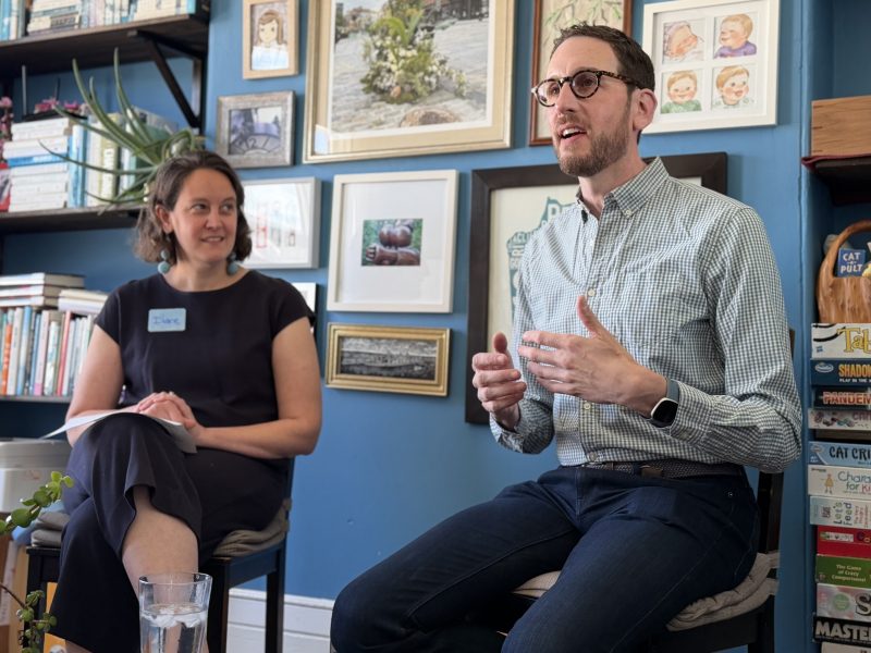 A man speaks while seated next to a woman taking notes in a room with bookshelves and framed art on a blue wall.