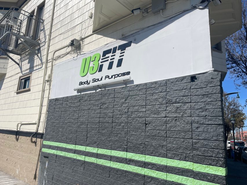 A gym sign reading "U3FIT Body Soul Purpose" is mounted on the corner of a gray and white building with green accent lines, in daytime.