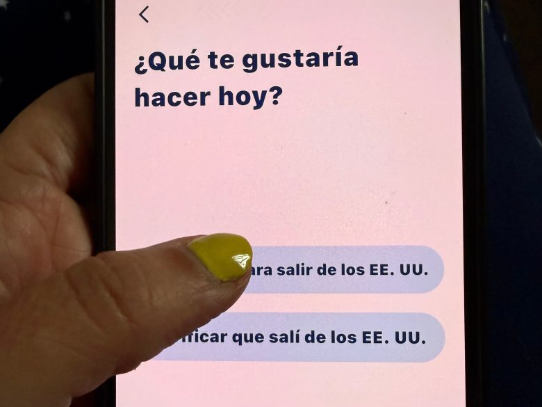 A hand with yellow nail polish holds a phone displaying a Spanish app for asylum-seekers that asks, "¿Qué te gustaría hacer hoy?" with two partially visible response options.