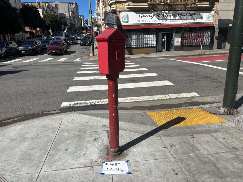 A red public mailbox on a street corner with a "WET PAINT" sign taped to the sidewalk below it; crosswalk and market visible in the background.