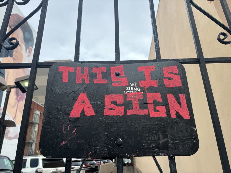 A black sign with the words "THIS IS A SIGN" in large red letters, and smaller text in the middle reading "WE SUING EVERYBODY," mounted on a metal gate.
