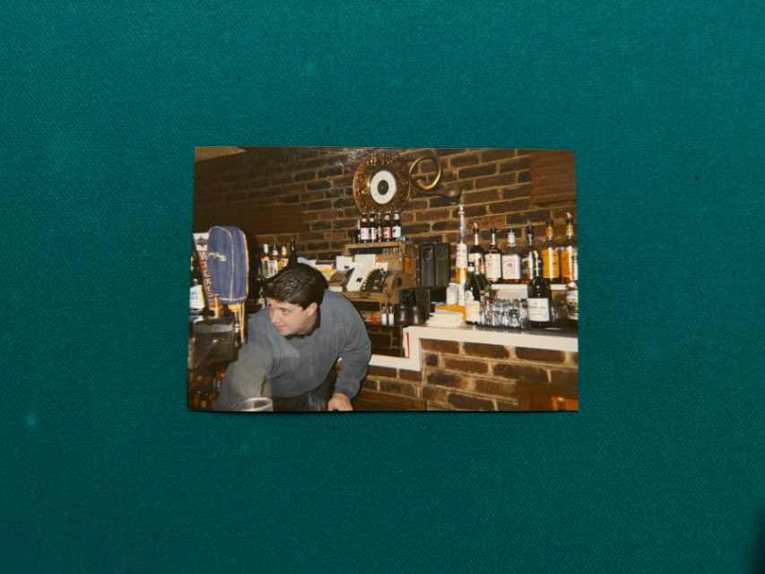 A man wearing glasses and a hat stands behind a bar with shelves of liquor bottles and a brick wall in the background.