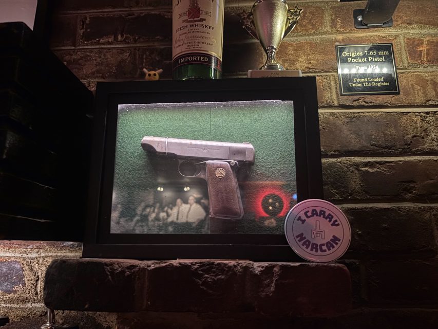 A framed 7.65 mm pocket pistol displayed on a green background, with a "I Carry Narcan" badge in front and bottles and plaques on a brick wall behind.