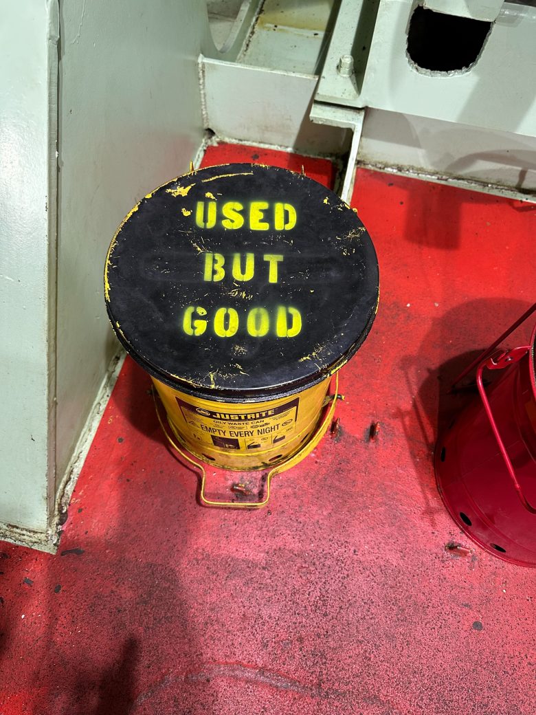 A yellow metal container with a black lid labeled "USED BUT GOOD" sits on a red floor next to industrial equipment.