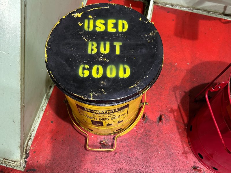 A yellow metal container with a black lid labeled "USED BUT GOOD" sits on a red floor next to industrial equipment.