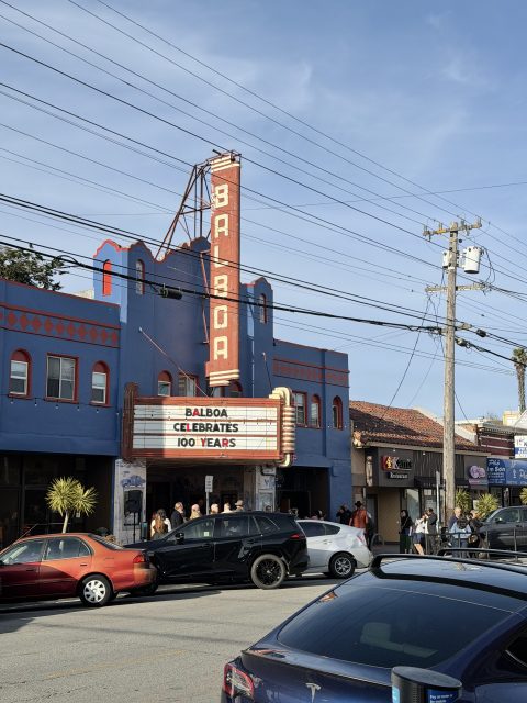 Street view of the Balboa Theatre with a marquee that reads "Balboa Celebrates 100 Years," cars parked in front, and people walking nearby.