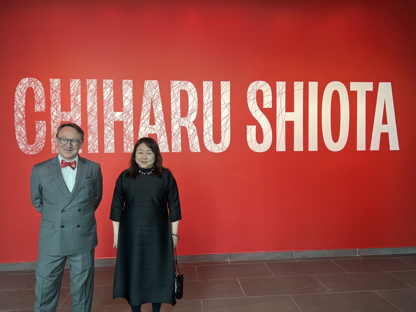 Two people stand in front of a red wall with large white text that reads "CHIHARU SHIOTA.
